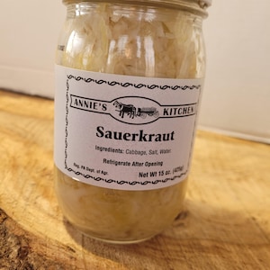May include: A glass jar of Annie's Kitchen Sauerkraut with a white lid. The label is white with black text and a black and white illustration of a horse-drawn wagon. The label reads "Annie's Kitchen Sauerkraut" and includes the ingredients: Cabbage, Salt, Water. It also states "Refrigerate After Opening" and "Net Wt 15 oz. (425g)".