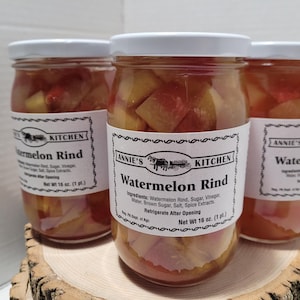 May include: Three glass jars filled with watermelon rind preserved in a clear liquid. The jars have white lids and labels that say "Annie's Kitchen" and "Watermelon Rind". The labels list the ingredients and instructions to refrigerate after opening. The jars are sitting on a wooden surface.