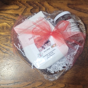 May include: A heart-shaped gift basket filled with a jar of strawberry preserves, a wrapped package, and decorative paper. The items are tied together with a red ribbon. The basket sits on a wooden surface.