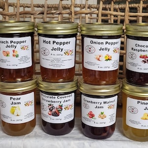 Op de afbeelding: Een verzameling glazen potten gevuld met verschillende soorten gelei, waaronder abrikoos-ananas, perzik-peper en chocolade-framboos. Elke pot heeft een gouden deksel en een etiket met de smaaknaam en een afbeelding. Elke pot bevat 227g gelei.
