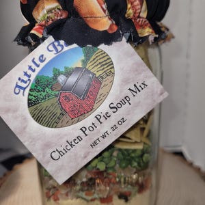 Puede incluir: Un tarro de vidrio lleno de mezcla para sopa de pollo y pastel, etiquetado "Little B" con una escena de granja. El tarro está cubierto con una tapa de tela negra con un diseño de perrito caliente. Peso neto 624 gramos.