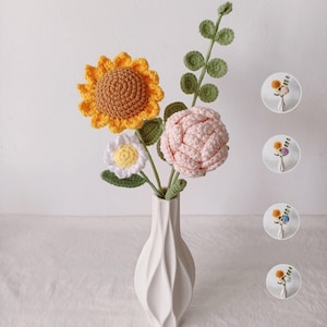 May include: A white ceramic vase holds a bouquet of handmade crochet flowers. The bouquet includes a yellow sunflower, a white daisy, and a pink peony. Green leaves and stems add detail to the arrangement. Small, circular images of the bouquet are in the background.