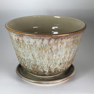 May include: A ceramic planter with a matching saucer featuring a textured exterior with a blend of light blue, cream, and metallic bronze glazes. The interior is a smooth, light beige finish with a dark brown rim along the top edge of the bowl.