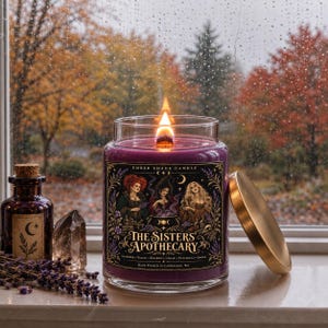 May include: A purple candle in a clear glass jar with a gold lid, featuring a label titled The Sisters' Apothecary with an illustration of three figures and a cauldron. The candle has a lit wooden wick and sits next to a small bottle and crystal.
