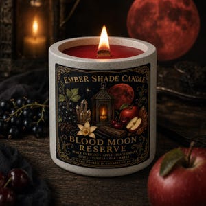 May include: A speckled white ceramic candle with a wooden wick and red wax. The label features the text Ember Shade Candle, Blood Moon Reserve, and lists black currant, apple, black cherry, clove, vanilla, oak, and amber as scent notes.