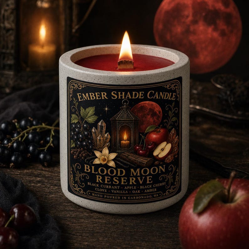 May include: A speckled white ceramic candle with a wooden wick and red wax. The label features the text Ember Shade Candle, Blood Moon Reserve, and lists black currant, apple, black cherry, clove, vanilla, oak, and amber as scent notes.