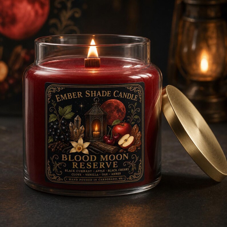 May include: A red wax candle in a clear glass jar with a wooden wick and a gold lid. The label reads Ember Shade Candle Blood Moon Reserve, featuring illustrations of a lantern, moon, fruit, and crystals, with text listing scent notes.
