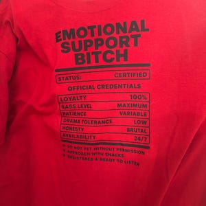 K&ouml;nnte beinhalten: Ein leuchtend rotes T-Shirt mit schwarzem Text, der EMOTIONAL SUPPORT BITCH lautet, gefolgt von einer Liste von Qualifikationen wie loyalty 100%, sass level maximum, patience variable, drama tolerance low, honesty brutal und availability 24/7.