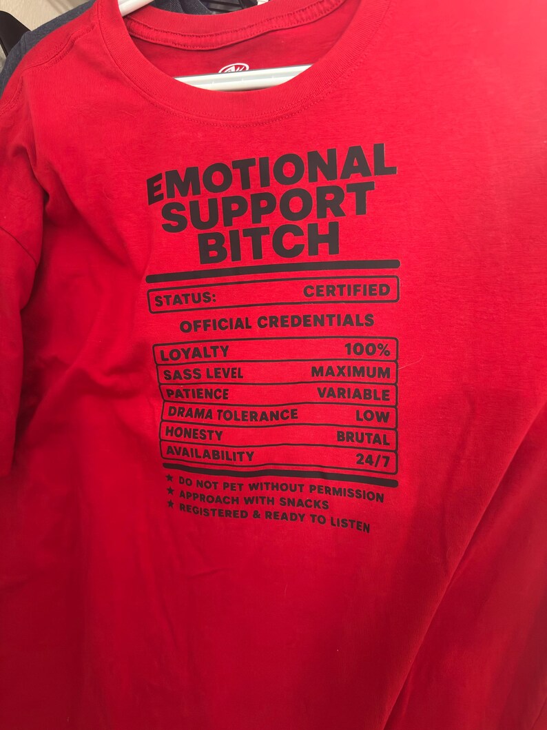 K&ouml;nnte beinhalten: Ein leuchtend rotes T-Shirt mit schwarzem Text, der EMOTIONAL SUPPORT BITCH lautet, gefolgt von einer Liste von Qualifikationen wie loyalty 100%, sass level maximum, patience variable, drama tolerance low, honesty brutal und availability 24/7.