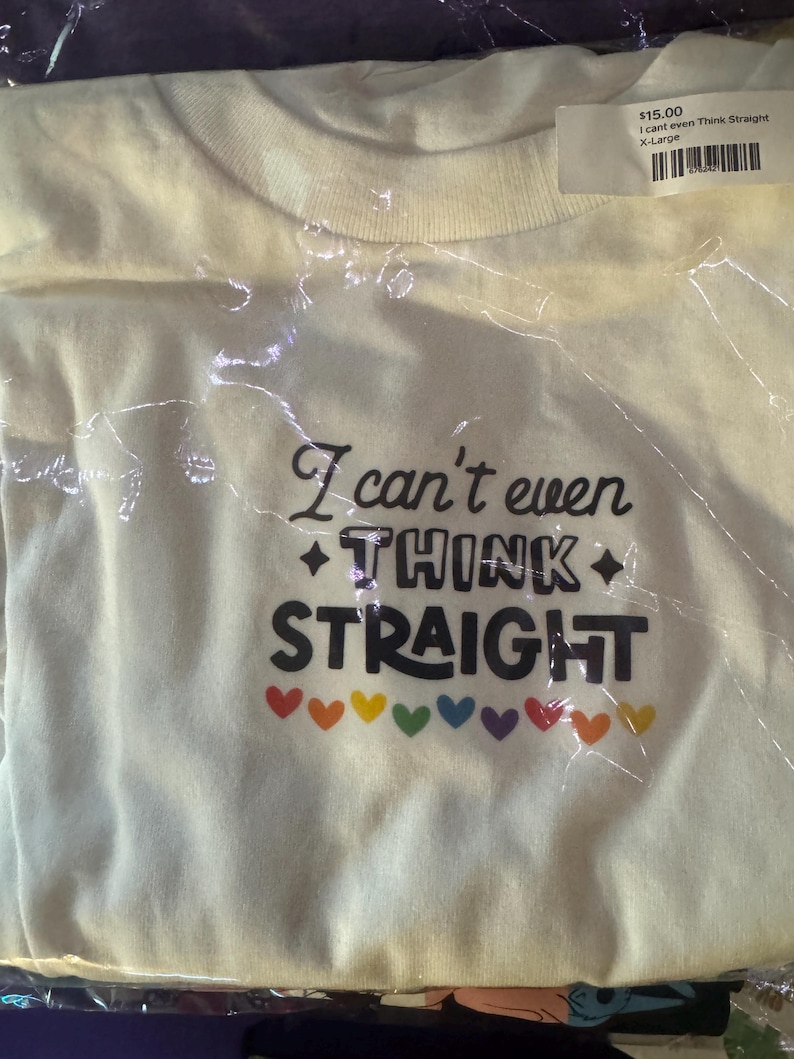 May include: A cream-coloured short-sleeve t-shirt featuring the black text "I can't even THINK STRAIGHT" with two black diamond symbols on either side of the word "THINK" and a row of small, colourful hearts in red, orange, yellow, green, blue, and purple.