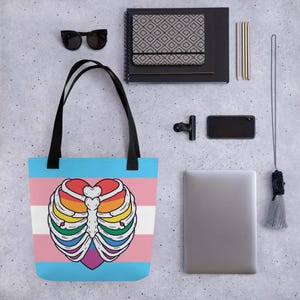 May include: A blue tote bag with black handles. The bag features a heart-shaped rib cage design in rainbow colours over a transgender flag. Other items include a black notebook and sunglasses.