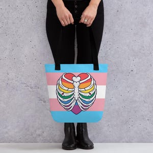 May include: A blue tote bag with a heart-shaped rib cage design in rainbow colours, set against a transgender pride flag. The bag has black handles. The bag is held by a person wearing black trousers and boots.