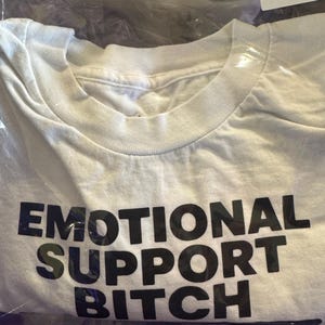 K&ouml;nnte beinhalten: Ein wei&szlig;es kurz&auml;rmeliges T-Shirt mit dem Text EMOTIONAL SUPPORT BITCH in fetten schwarzen Gro&szlig;buchstaben auf der Brust. Das Kleidungsst&uuml;ck ist in einer durchsichtigen Plastikt&uuml;te verpackt, an deren oberer rechter Ecke ein wei&szlig;es Etikett klebt.