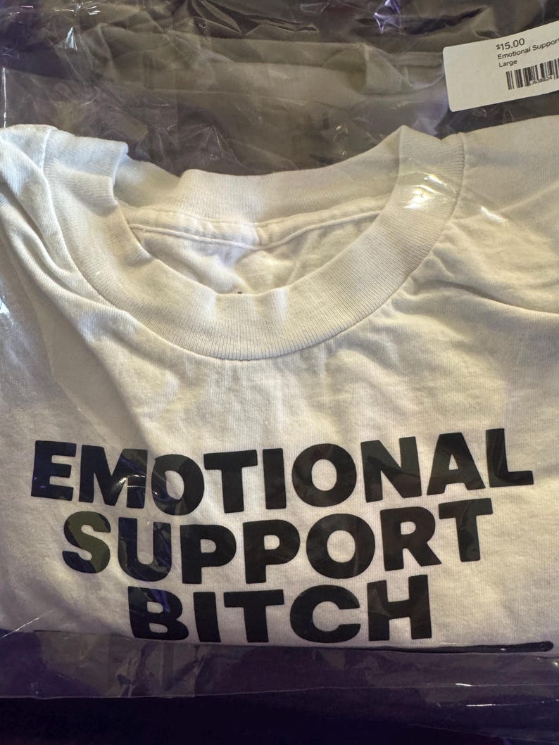 K&ouml;nnte beinhalten: Ein wei&szlig;es kurz&auml;rmeliges T-Shirt mit dem Text EMOTIONAL SUPPORT BITCH in fetten schwarzen Gro&szlig;buchstaben auf der Brust. Das Kleidungsst&uuml;ck ist in einer durchsichtigen Plastikt&uuml;te verpackt, an deren oberer rechter Ecke ein wei&szlig;es Etikett klebt.