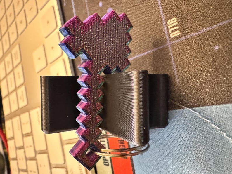 K&ouml;nnte beinhalten: Ein pixeliger Axt-Schl&uuml;sselanh&auml;nger mit einer texturierten, irisierenden violetten und schwarzen Oberfl&auml;che. Der Axtkopf hat ein blockartiges, gezacktes Design und der Griff verf&uuml;gt &uuml;ber einen runden Metallring an der Basis f&uuml;r Schl&uuml;ssel.