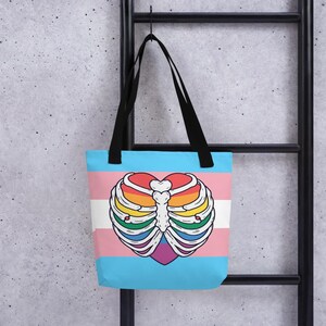 May include: A blue tote bag with black handles, displaying a heart-shaped rib cage design in rainbow hues. The bag incorporates the transgender pride flag colours: pink, white, and light blue. The bag is suspended against a grey background.