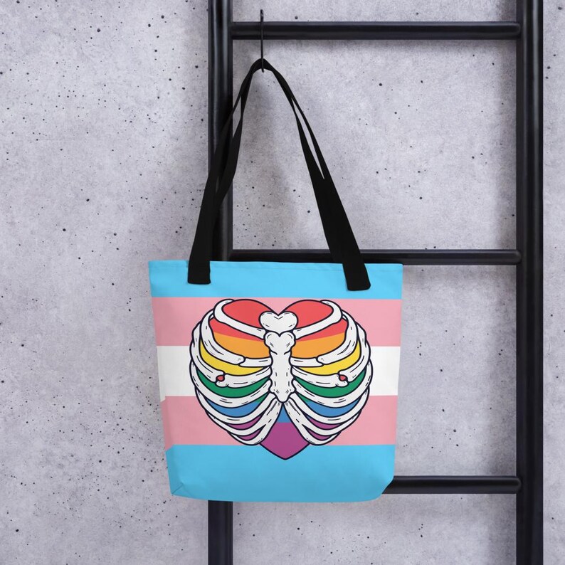 May include: A blue tote bag with black handles, displaying a heart-shaped rib cage design in rainbow hues. The bag incorporates the transgender pride flag colours: pink, white, and light blue. The bag is suspended against a grey background.