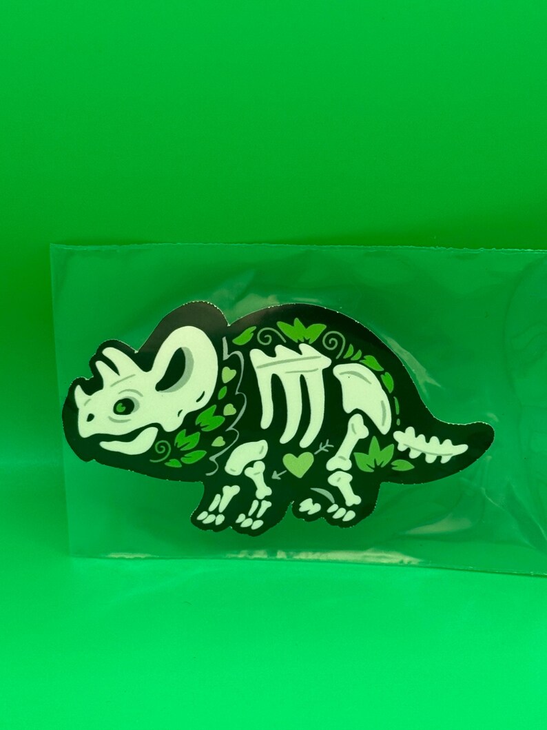 May include: A sticker featuring a stylized triceratops skeleton in pale green against a black background. The design incorporates decorative leaf patterns and small heart shapes integrated into the dinosaur's body, all enclosed in a clear plastic sleeve.