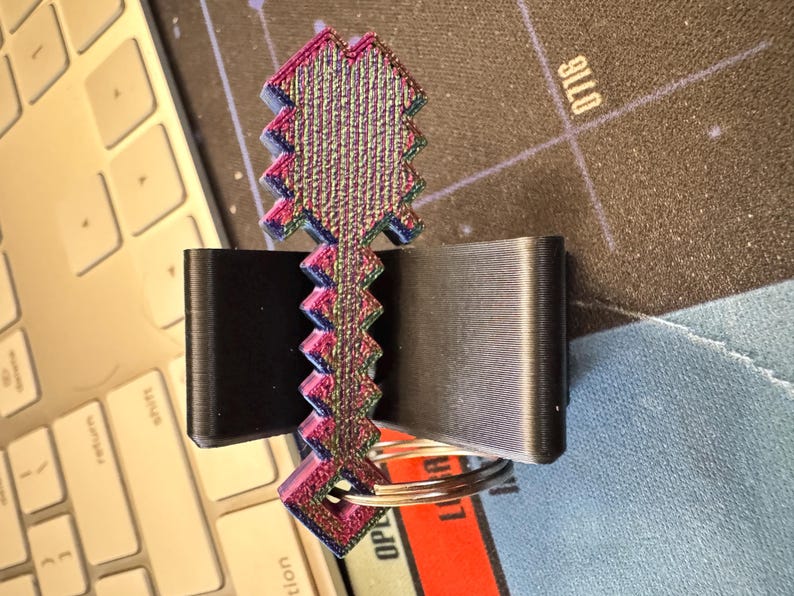 May include: A 3D-printed keychain featuring a pixelated sword design with a vibrant, iridescent color gradient shifting between purple, blue, and green. The sword has a jagged, blocky silhouette and includes a circular metal split ring at the base.