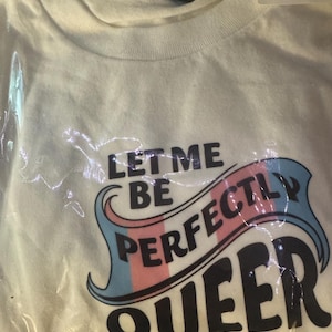 May include: A white short-sleeved t-shirt packaged in clear plastic. The shirt features the black text "LET ME BE PERFECTLY QUEER" with the word "PERFECTLY" overlaid on a stylised banner graphic coloured in light blue, pink, and white stripes.