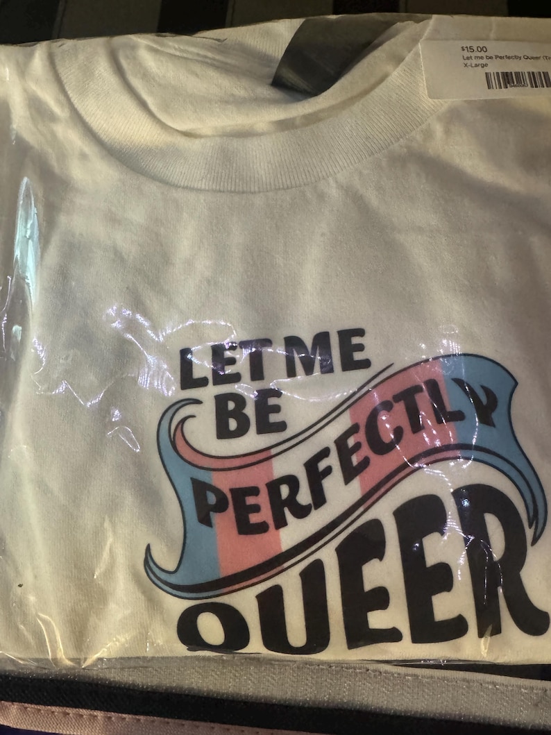 May include: A white short-sleeved t-shirt packaged in clear plastic. The shirt features the black text "LET ME BE PERFECTLY QUEER" with the word "PERFECTLY" overlaid on a stylised banner graphic coloured in light blue, pink, and white stripes.