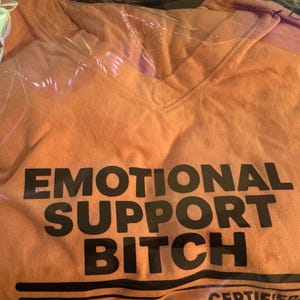 K&ouml;nnte beinhalten: Ein orangefarbenes T-Shirt mit V-Ausschnitt und dem schwarzen Schriftzug EMOTIONAL SUPPORT BITCH in fetter serifenloser Schrift auf der Brust, sowie weiterem schwarzem Text und horizontalen Linien am unteren Rand in transparenter Verpackung.