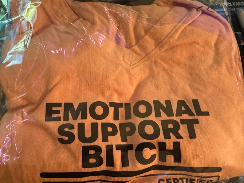 K&ouml;nnte beinhalten: Ein orangefarbenes T-Shirt mit V-Ausschnitt und dem schwarzen Schriftzug EMOTIONAL SUPPORT BITCH in fetter serifenloser Schrift auf der Brust, sowie weiterem schwarzem Text und horizontalen Linien am unteren Rand in transparenter Verpackung.