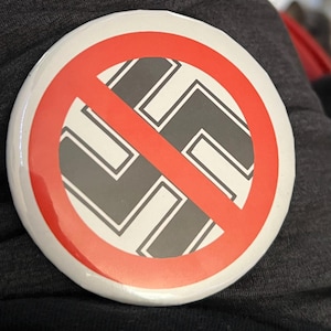 May include: A circular pinback button featuring a white background with a black swastika symbol centred in the middle. A bold red diagonal slash crosses over the swastika from the upper left to the lower right, indicating a prohibition symbol.