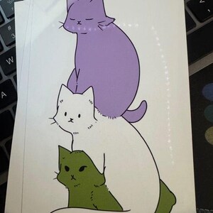 May include: A rectangular sticker featuring a vertical stack of three illustrated cats. The top cat is light purple with closed eyes, the middle cat is white with black eyes, and the bottom cat is olive green with a focused expression and black outlines.