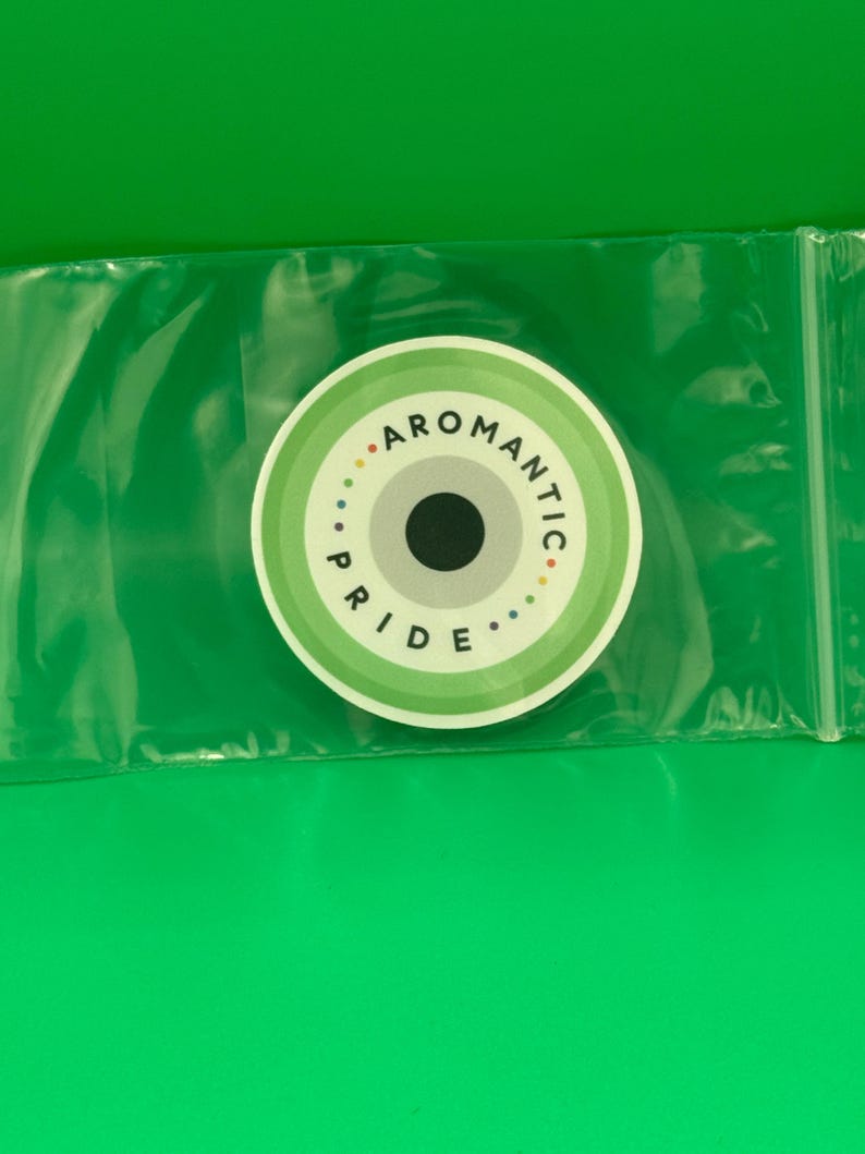 Op de afbeelding: Een ronde sticker met concentrische ringen in tinten groen, wit en grijs. Het midden bevat een effen zwarte cirkel. De tekst AROMANTIC PRIDE is in het zwart gedrukt langs de bovenste en onderste boog, geaccentueerd door kleine gekleurde stippen.