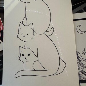 May include: A white rectangular card featuring a black ink line drawing of three cats stacked vertically. The top cat has closed eyes, the middle cat has open eyes, and the bottom cat has a small face, all depicted in a simple, clean illustration style.
