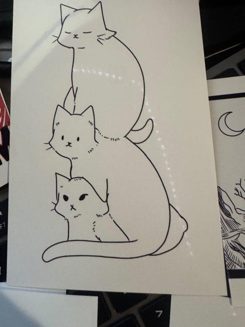 May include: A white rectangular card featuring a black ink line drawing of three cats stacked vertically. The top cat has closed eyes, the middle cat has open eyes, and the bottom cat has a small face, all depicted in a simple, clean illustration style.