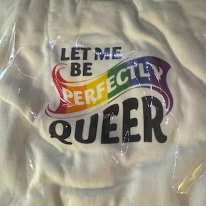 May include: A white t-shirt packaged in clear plastic, featuring the black text "LET ME BE" above the word "PERFECTLY" in a rainbow-coloured font, followed by the black text "QUEER" in a bold, rounded typeface on the front of the garment.