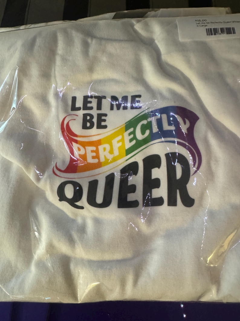 May include: A white t-shirt packaged in clear plastic, featuring the black text "LET ME BE" above the word "PERFECTLY" in a rainbow-coloured font, followed by the black text "QUEER" in a bold, rounded typeface on the front of the garment.