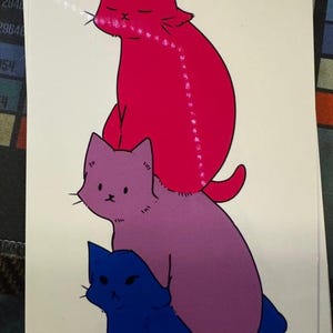 May include: A vertical illustration on a white background featuring three stacked cats. The top cat is bright pink, the middle cat is purple, and the bottom cat is dark blue. Each cat has simple facial features and a solid colour fill.