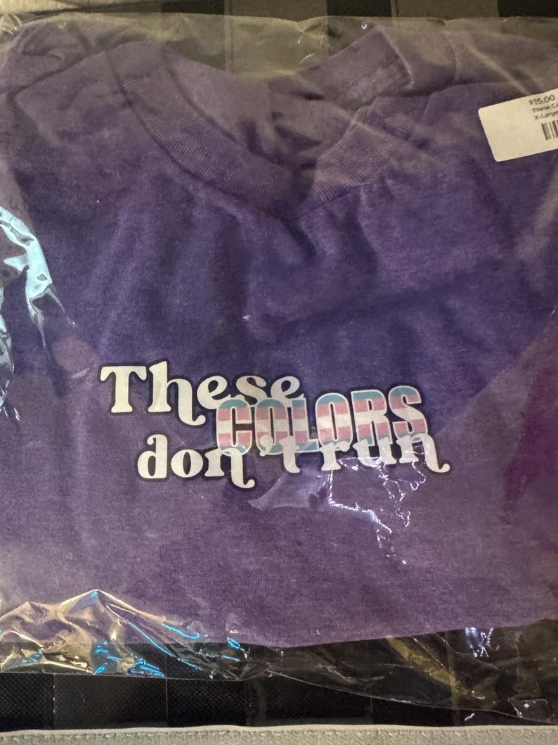 May include: A purple short-sleeved t-shirt packaged in a clear plastic bag. The shirt features the white text "These" and "don't run" with the word "COLORS" in the centre, which is filled with a light blue, pink, and white striped pattern.