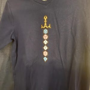 Op de afbeelding: Een marineblauw T-shirt met V-hals voorzien van een grafische print van een zwaard bestaande uit gestapelde veelzijdige dobbelstenen in blauwe, roze en witte tinten. Het gevest van het zwaard is goudkleurig. Het label in de nek toont de tekst [g].