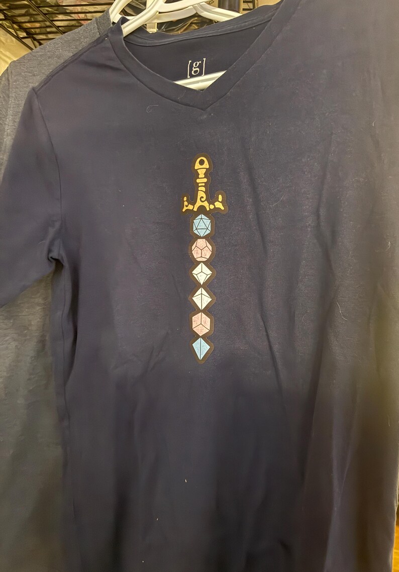 Op de afbeelding: Een marineblauw T-shirt met V-hals voorzien van een grafische print van een zwaard bestaande uit gestapelde veelzijdige dobbelstenen in blauwe, roze en witte tinten. Het gevest van het zwaard is goudkleurig. Het label in de nek toont de tekst [g].