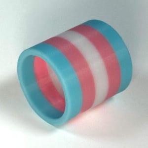 Op de afbeelding: Een cilindrische ring met een ontwerp met afwisselende banden van lichtblauw, roze en wit. Het ontwerp van de ring vertegenwoordigt de transgender pride vlag. De ring is gemaakt van een glad materiaal.