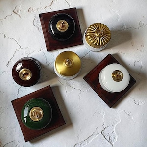 May include: A collection of vintage-style light switches in various colors, including black, white, green, and brown. Each switch has a gold-colored toggle and is mounted on a dark wooden square. The switches are arranged on a textured, off-white surface.