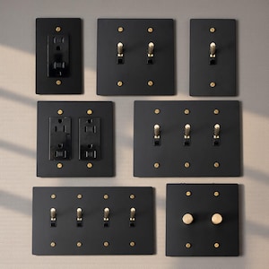 Può includere: Una collezione di placche elettriche nere con dettagli color ottone. Le placche includono prese di corrente, interruttori singoli e doppi e dimmer. Le placche sono montate su una parete grigio chiaro.