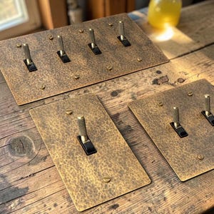 May include: Several antique-style light switch plates with a hammered brass finish. Each plate has a black toggle switch and brass screws. The plates are rectangular and vary in size, with one holding four switches.