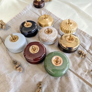 Könnte beinhalten: Eine Sammlung von keramikartigen Schaltern in verschiedenen Farben, darunter Schwarz, Weiß, Gold und Grün. Jede Box hat einen goldfarbenen Schalter und steht auf einer neutralfarbenen Oberfläche. Die Boxen sind rund und haben eine glänzende Oberfläche.