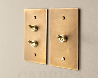 May include: Two rectangular brass light switch plates with toggle switches. Each plate has a brushed finish and is mounted on a neutral-colored wall. The switches have a classic, vintage design.