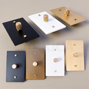 May include: A collection of rectangular light switch plates in various colors, including black, white, gold, and bronze. Each plate features a textured gold knob and matching screws, showcasing a modern design aesthetic.