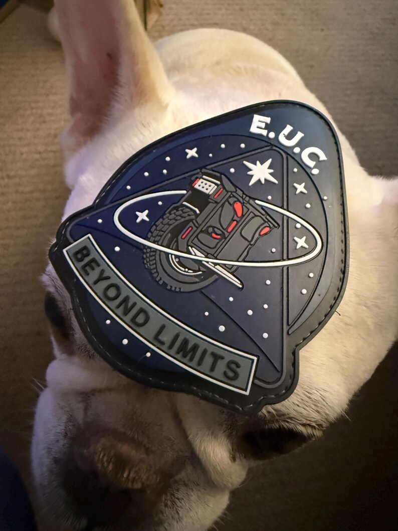 Pode incluir: Um patch azul marinho com o texto "BEYOND LIMITS" e "E.U.C." apresenta um gr&aacute;fico de um ve&iacute;culo. O patch est&aacute; definido contra o fundo de um c&atilde;o de cor clara.