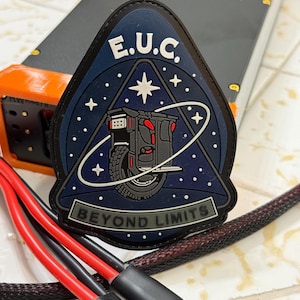 Pode incluir: Um patch azul escuro, preto e cinza com o texto "E.U.C." e "BEYOND LIMITS". O patch apresenta um gr&aacute;fico de um ve&iacute;culo de uma roda contra um fundo estrelado. Fios vermelhos e pretos em primeiro plano.