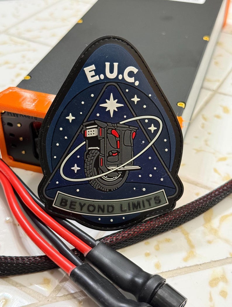 Pode incluir: Um patch azul escuro, preto e cinza com o texto "E.U.C." e "BEYOND LIMITS". O patch apresenta um gr&aacute;fico de um ve&iacute;culo de uma roda contra um fundo estrelado. Fios vermelhos e pretos em primeiro plano.
