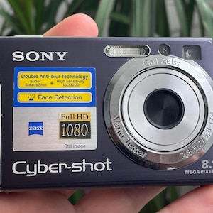 May include: A dark blue Sony Cyber-shot digital camera with a silver lens and accents. Features include Full HD 1080, 8.1 megapixels, and Carl Zeiss optics. Includes text: Double Anti-blur Technology, Face Detection.
