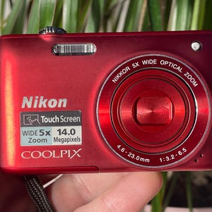 May include: A red Nikon Coolpix digital camera with a 5x wide optical zoom lens. Features include a touch screen and 14.0 megapixels. The lens has a focal length of 4.6-23.0mm and an aperture of 1:3.2-6.5.